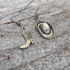 May include: Silver-toned cowboy-themed earrings. One earring features a cowboy boot charm, the other a cowboy hat charm. The charms are detailed with intricate designs and hang from silver-colored ear wires. The earrings are displayed on a weathered wooden surface.