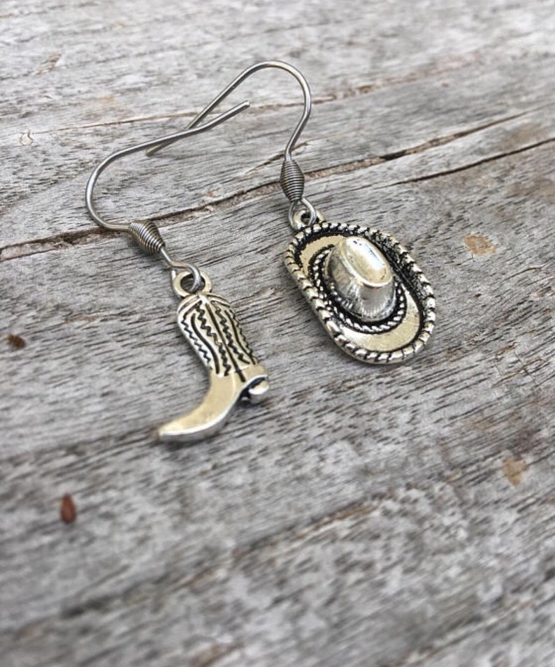 May include: Silver-toned cowboy-themed earrings. One earring features a cowboy boot charm, the other a cowboy hat charm. The charms are detailed with intricate designs and hang from silver-colored ear wires. The earrings are displayed on a weathered wooden surface.