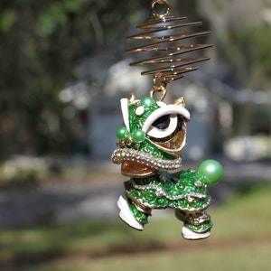 May include: A decorative ornament featuring a green and gold lion dance figure. The lion has white accents, a jeweled collar, and green pearl-like embellishments. It hangs from a gold wire spiral.