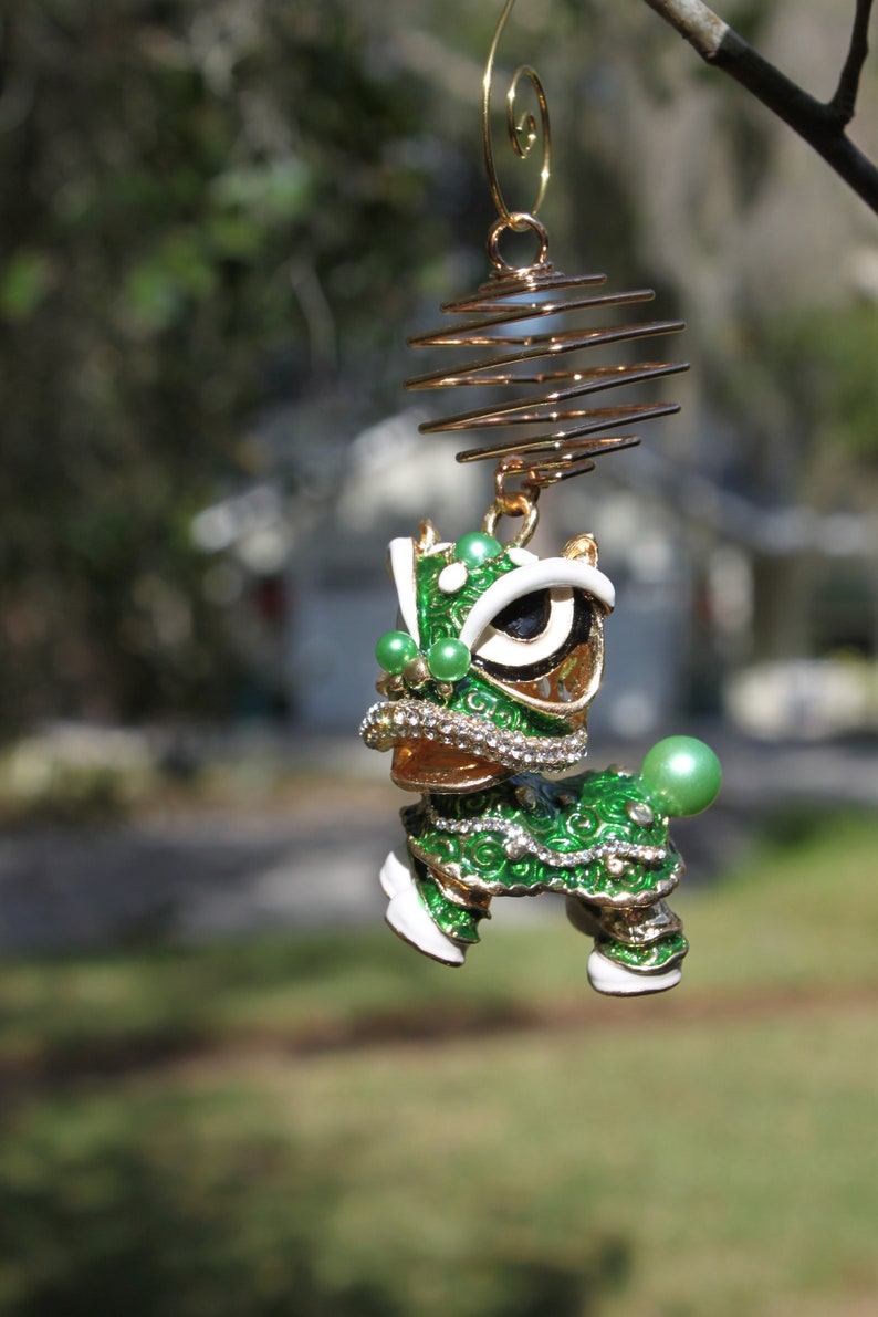 May include: A decorative ornament featuring a green and gold lion dance figure. The lion has white accents, a jeweled collar, and green pearl-like embellishments. It hangs from a gold wire spiral.