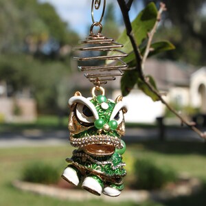 May include: A decorative ornament featuring a green and gold lion dance figure with white accents and sparkling embellishments. The ornament is suspended from a gold-colored spiral and hook, hanging from a tree branch.
