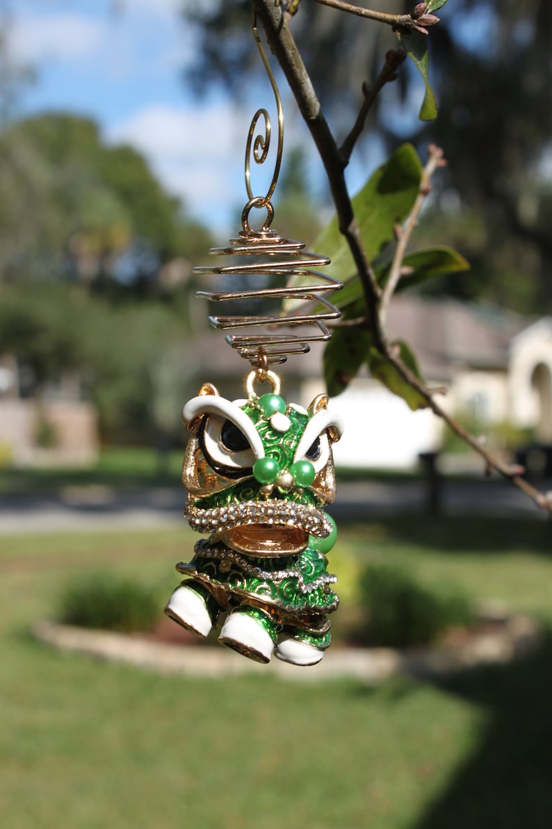 May include: A decorative ornament featuring a green and gold lion dance figure with white accents and sparkling embellishments. The ornament is suspended from a gold-colored spiral and hook, hanging from a tree branch.