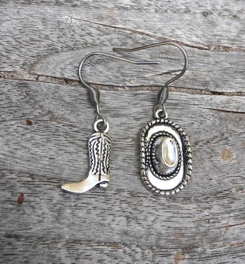 May include: Silver-toned earrings featuring a cowboy boot and a cowboy hat charm. The boot has detailed stitching, and the hat has a rope-like border. The earrings hang from a simple hook.