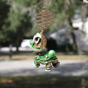 May include: A decorative, jeweled turtle ornament with a gold-colored wire spiral and cage. The turtle is green, white, and gold with rhinestone accents and a large green pearl on its back. The ornament hangs from a tree branch.