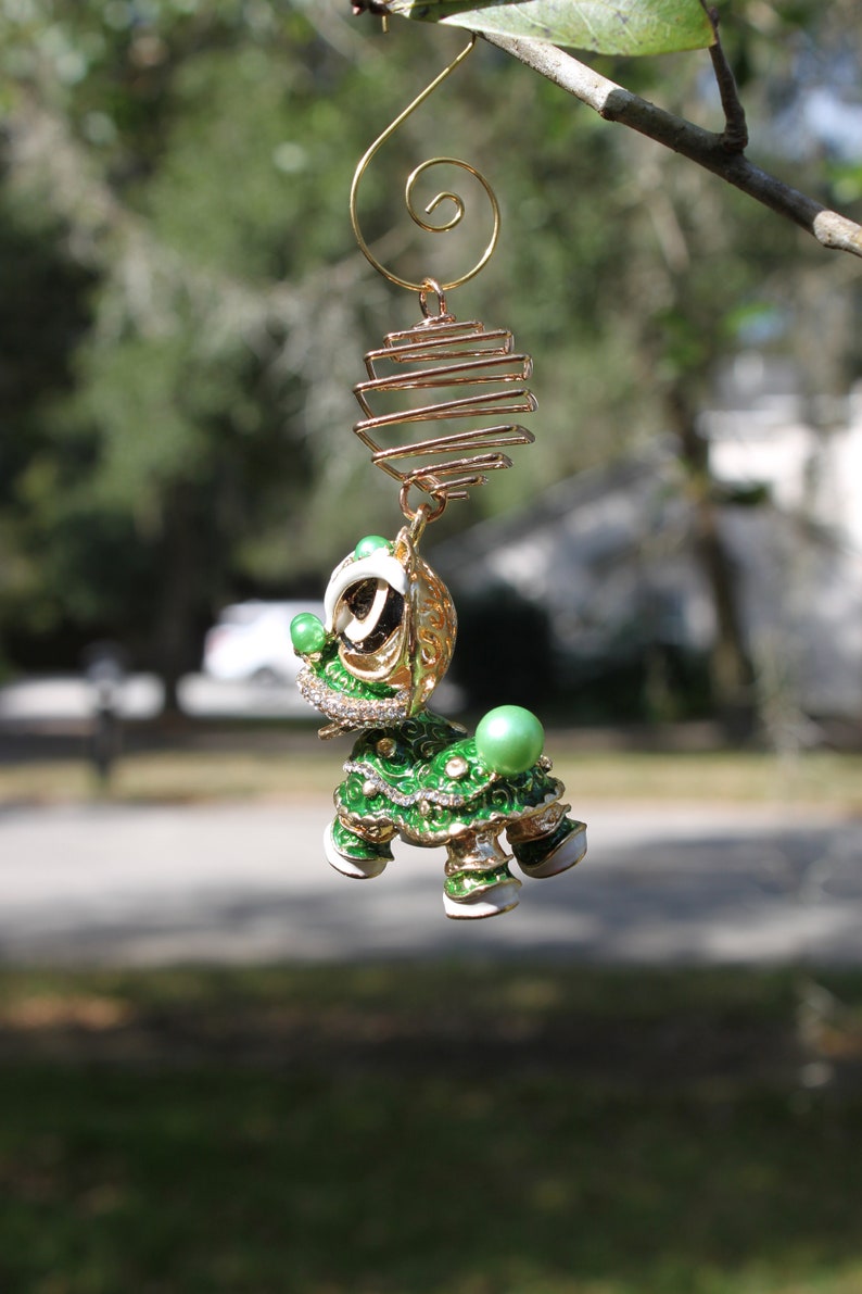 May include: A decorative, jeweled turtle ornament with a gold-colored wire spiral and cage. The turtle is green, white, and gold with rhinestone accents and a large green pearl on its back. The ornament hangs from a tree branch.