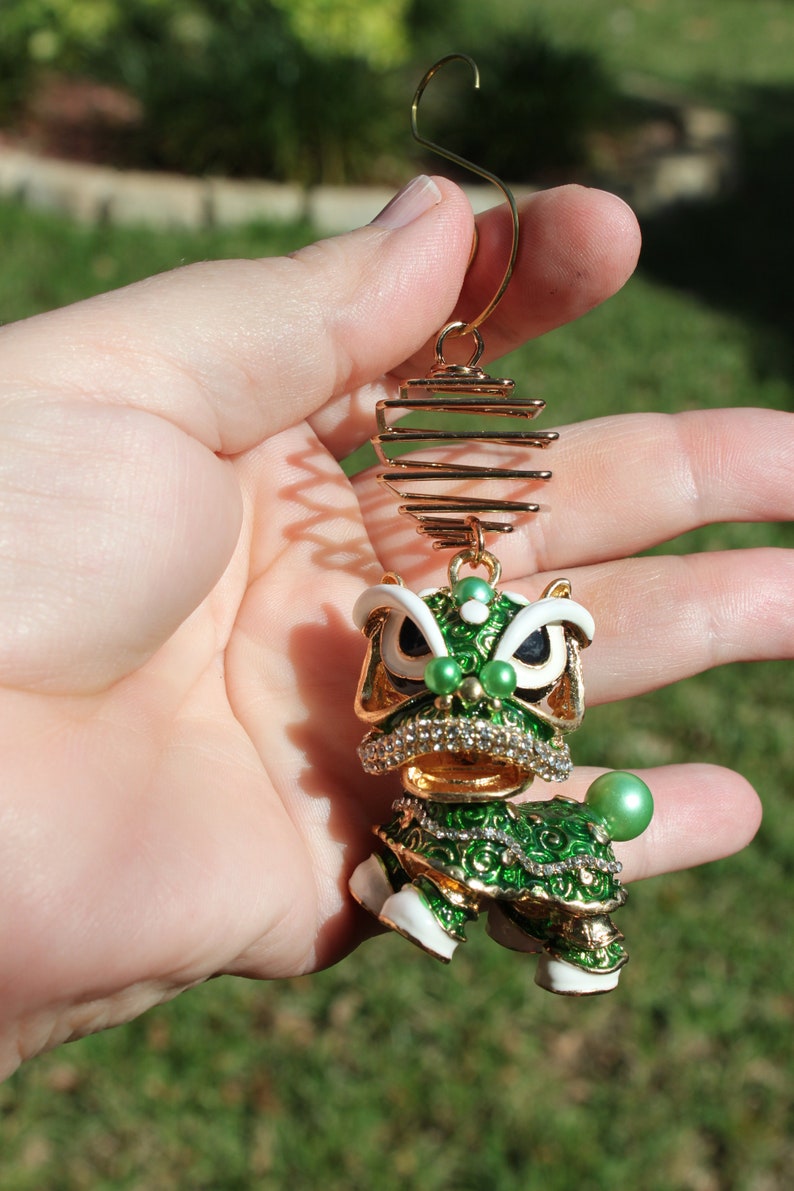 May include: A gold-toned ornament featuring a detailed green and white lion dance figure. The lion has a jeweled collar and is accented with pearls. The ornament is suspended from a gold wire.