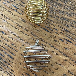 May include: Two wire-wrapped pendants. The top pendant is gold-colored, spherical, and spiral-shaped. The bottom pendant is silver-colored, with a geometric, cage-like design. Both pendants have a small loop at the top for hanging.