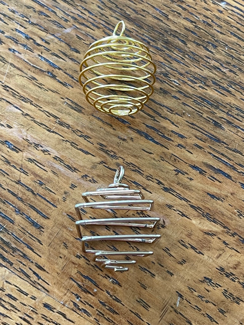 May include: Two wire-wrapped pendants. The top pendant is gold-colored, spherical, and spiral-shaped. The bottom pendant is silver-colored, with a geometric, cage-like design. Both pendants have a small loop at the top for hanging.