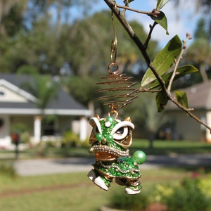 May include: A gold-toned earring featuring a green and white lion dance figure with intricate details. The lion has a detailed face, embellished with rhinestones and green beads. The earring is suspended from a gold spiral and hook.