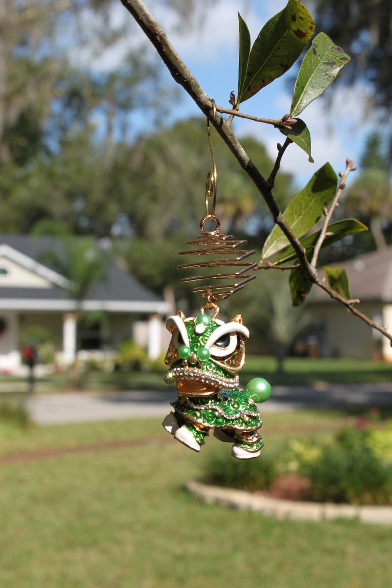 May include: A gold-toned earring featuring a green and white lion dance figure with intricate details. The lion has a detailed face, embellished with rhinestones and green beads. The earring is suspended from a gold spiral and hook.