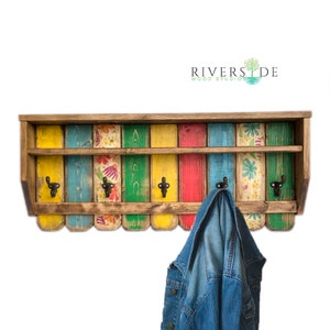 May include: A colorful wooden wall-mounted coat rack with a shelf. The rack is painted in shades of yellow, green, blue, red, and brown. It has five hooks and a floral design on the front. The rack is made of reclaimed wood and has a rustic look. "Riverside Wood Studio" is printed on the top of the rack.
