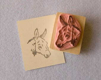 May include: A wooden mounted rubber stamp with a pink donkey head design. The stamp is used to create an impression of a donkey head on paper.