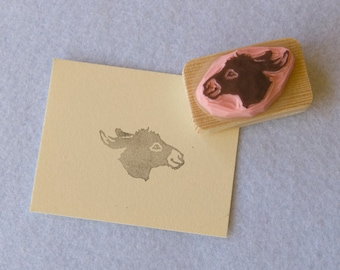 May include: A wooden block with a pink rubber stamp of a donkey's head. The stamp is on a light brown surface and a gray donkey head is stamped on the surface.