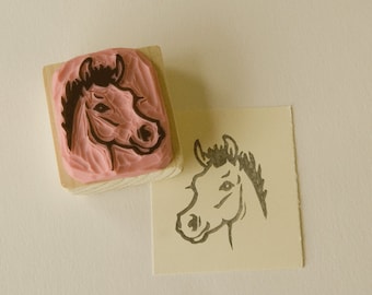 May include: A wooden stamp with a pink rubber surface featuring a horse head design. The stamp is next to a piece of paper with a black ink impression of the horse head.