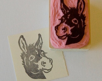 May include: A black and white rubber stamp with a donkey head design. The stamp is mounted on a wooden block with a pink rubber surface.