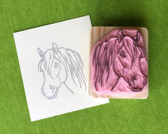 May include: A pink rubber stamp of a horse's head on a wooden block. A gray ink print of the same horse head is on a white card. The background is a textured green fabric.
