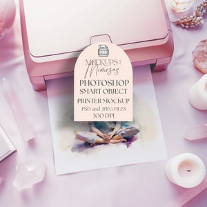 May include: A pink printer with a white label that says "Mockups and Mimosa's" and "Photoshop Smart Object Printer Mockup PSD and JPEG Files 300 DPI". The printer is on a pink surface with a watercolour painting of a person sitting with their legs crossed.