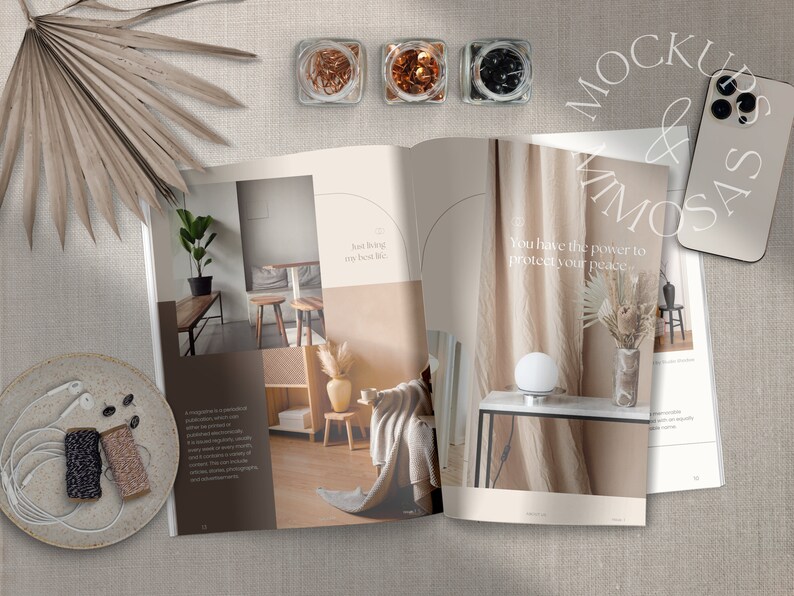 May include: Open magazine with interior design photos and text that reads "Just living my best life" and "You have the power to protect your peace."  The magazine is on a beige linen surface with a palm leaf, a plate with a pair of earbuds and two spools of twine, and a white smartphone.