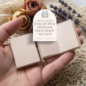May include: A miniature blank book mockup, held in a hand, with a soft beige cover and pages. The book is open, revealing the interior pages. The background includes dried roses and a label that reads "VINTAGE ROSES JUNK JOURNAL MINI BOOK PHOTOSHOP MOCKUP".