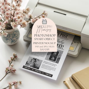 Puede incluir: Una maqueta de impresora con un fondo blanco y una hoja de papel que sale de la impresora. El papel tiene una foto en blanco y negro de una ecografía de bebé y el texto "Nuestro pequeño se espera que llegue en enero de 2026." El texto "Próximamente" está en el papel. La impresora es una impresora de inyección de tinta blanca con una bandeja negra y dorada. Hay un arreglo de flores rosas y blancas en un jarrón azul y blanco en la esquina superior izquierda de la imagen.