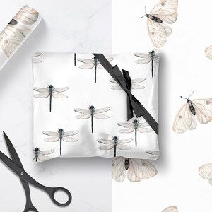 May include: White wrapping paper with a repeating pattern of delicate brown dragonflies and butterflies. A black ribbon is tied around the paper, and a pair of black scissors are lying on a white surface.