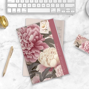 May include: A pink and white floral patterned gratitude journal with the text "Gratitude Journal" printed on the spine. The journal is lying on a white marble surface with a gold pen, a candle, and a small floral patterned card.