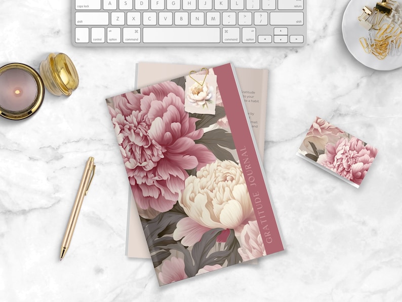 May include: A pink and white floral patterned gratitude journal with the text "Gratitude Journal" printed on the spine. The journal is lying on a white marble surface with a gold pen, a candle, and a small floral patterned card.