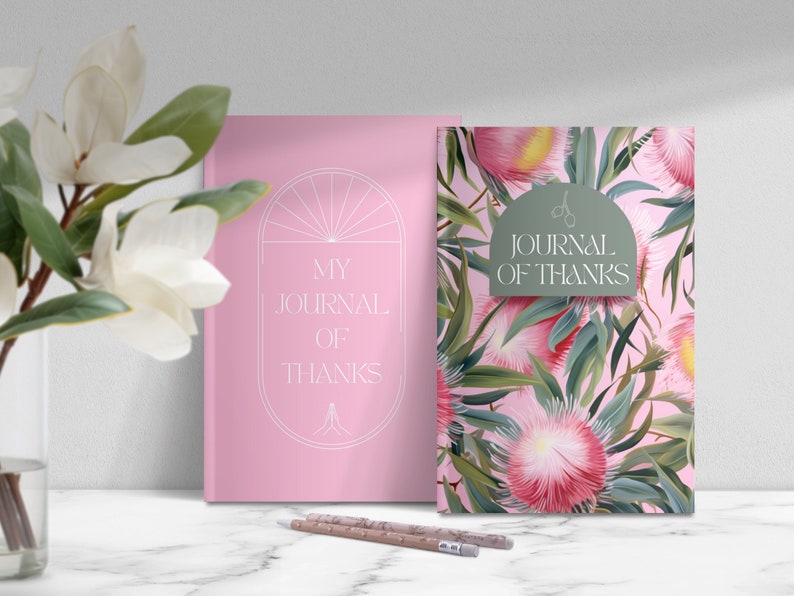 May include: Two pink journals with the text "My Journal of Thanks" on the front. One journal has a simple design with a circle and lines. The other journal has a floral pattern with pink flowers and green leaves.