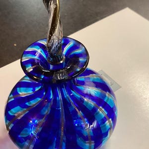 May include: A round glass perfume bottle featuring vibrant cobalt blue and turquoise vertical stripes with thin silver accents. The bottle has a wide, flat neck and a clear, twisted glass stopper that extends upwards in a spiral shape.