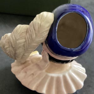 May include: A porcelain figurine of a woman wearing a blue hat with a white feather and a ruffled collar. The figurine has a light pink and cream colour palette, with a dark brown interior in the hat.