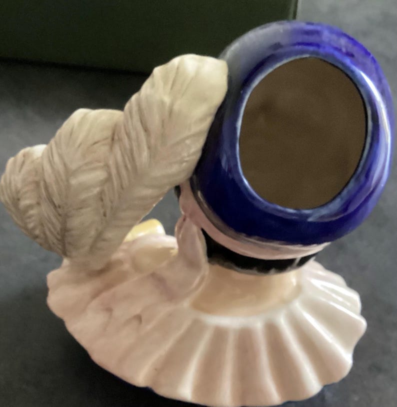 May include: A porcelain figurine of a woman wearing a blue hat with a white feather and a ruffled collar. The figurine has a light pink and cream colour palette, with a dark brown interior in the hat.