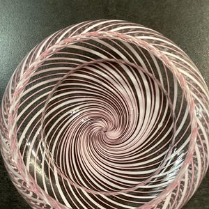 May include: A clear glass bowl featuring a swirling pattern of opaque white and soft pink lines that radiate from the center point toward the rim, creating a continuous spiral effect throughout the entire interior and exterior surface of the vessel.