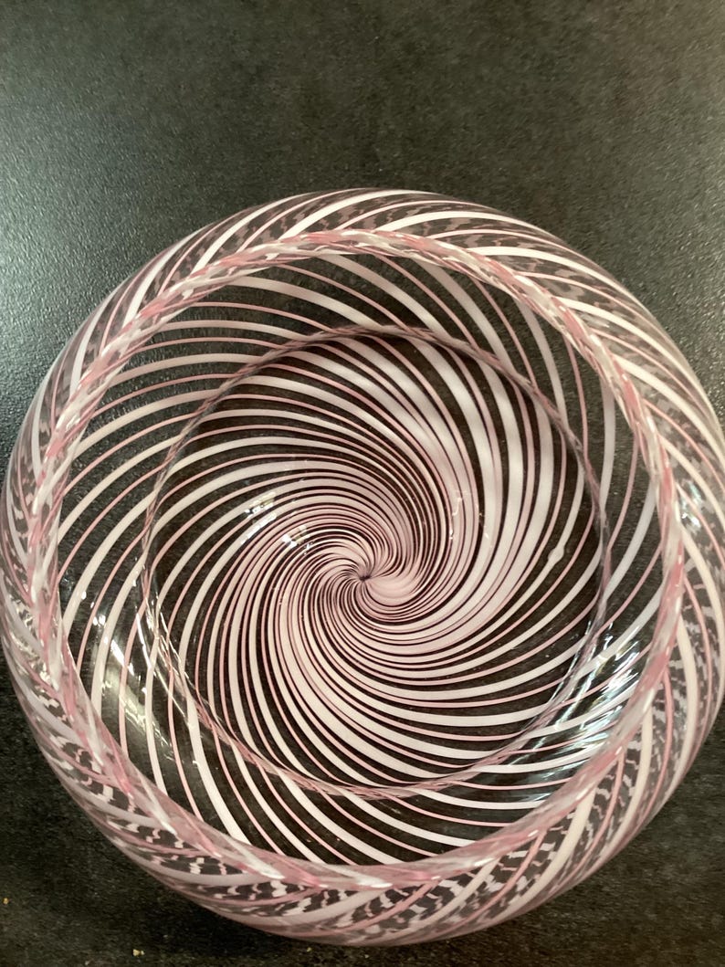 May include: A clear glass bowl featuring a swirling pattern of opaque white and soft pink lines that radiate from the center point toward the rim, creating a continuous spiral effect throughout the entire interior and exterior surface of the vessel.