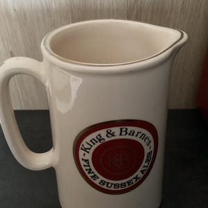 Op de afbeelding: Cr&egrave;mekleurige keramische kan met een handvat en een schenktuit. De kan heeft een cirkelvormig embleem met de tekst "King & Barnes FINE SUSSEX ALES" in een rood en goudkleurig ontwerp.