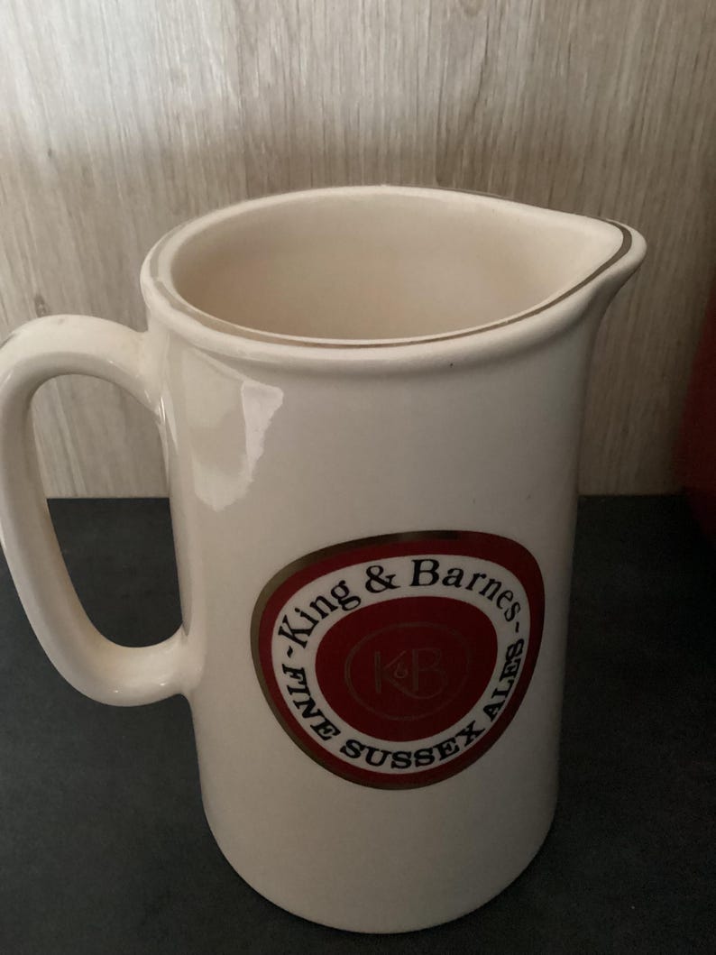 Op de afbeelding: Cr&egrave;mekleurige keramische kan met een handvat en een schenktuit. De kan heeft een cirkelvormig embleem met de tekst "King & Barnes FINE SUSSEX ALES" in een rood en goudkleurig ontwerp.