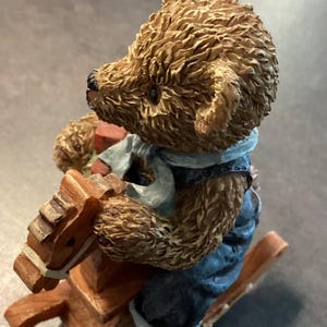 Può includere: Una statuina di orsacchiotto marrone che cavalca un cavallo a dondolo di legno. L'orso indossa una tuta blu e una sciarpa azzurra. L'orso tiene in mano un piccolo pacco rosso e marrone. La statuina ha un aspetto dettagliato e strutturato.
