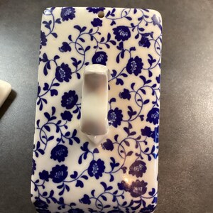 May include: A rectangular ceramic butter dish lid featuring a white base with a repeating blue floral and vine pattern. The lid includes a central white arched handle and a small circular hole near the top edge, showcasing a classic glazed finish.