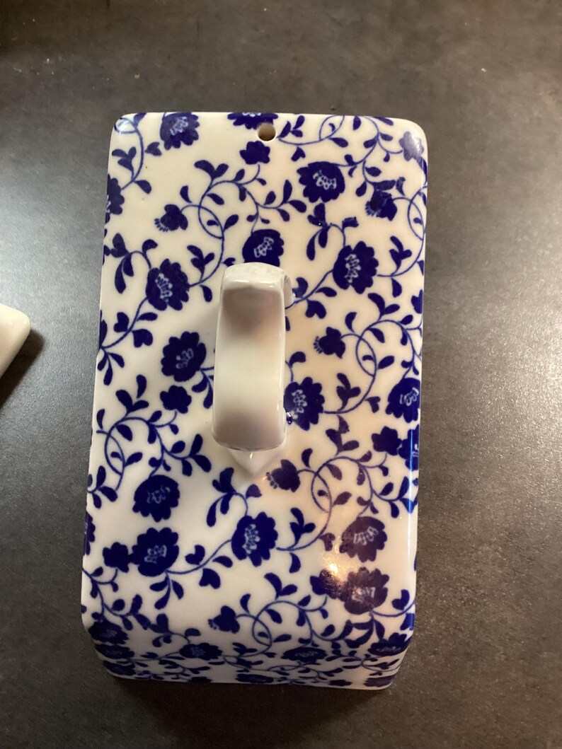 May include: A rectangular ceramic butter dish lid featuring a white base with a repeating blue floral and vine pattern. The lid includes a central white arched handle and a small circular hole near the top edge, showcasing a classic glazed finish.