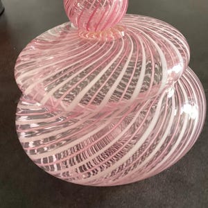 May include: A round glass jar with a matching lid, featuring a swirling pattern of translucent pink and opaque white stripes. The lid is topped with a spherical glass finial that mirrors the same pink and white spiral design as the main body.