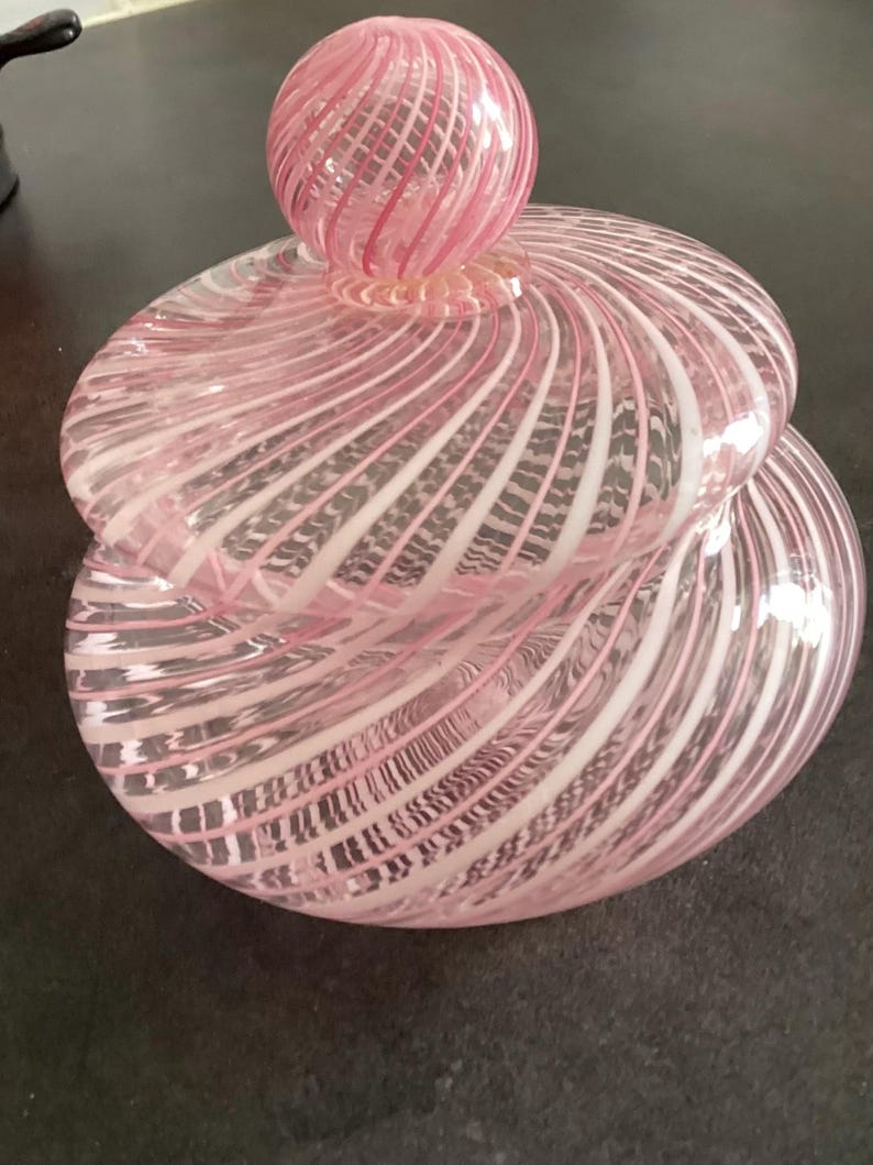 May include: A round glass jar with a matching lid, featuring a swirling pattern of translucent pink and opaque white stripes. The lid is topped with a spherical glass finial that mirrors the same pink and white spiral design as the main body.
