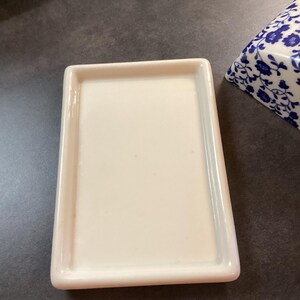 May include: A rectangular, off-white ceramic butter dish base with a smooth, flat surface and raised edges. Beside it sits a portion of a matching ceramic lid featuring a blue and white floral pattern, showcasing a classic, clean design.