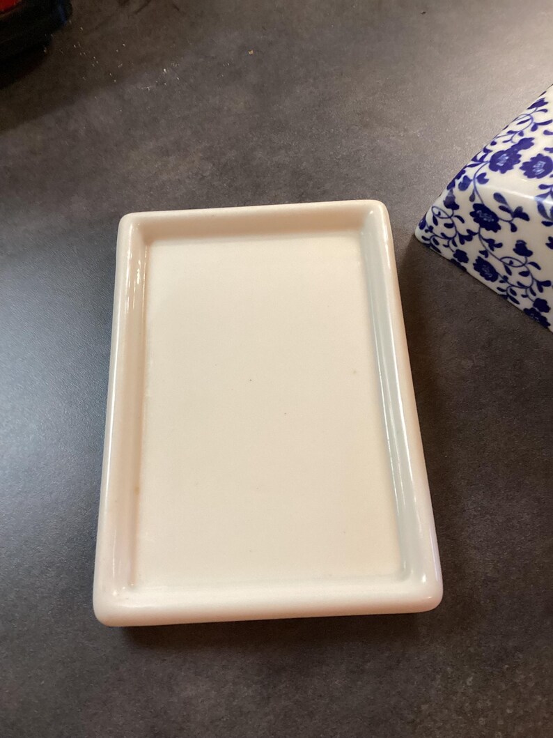 May include: A rectangular, off-white ceramic butter dish base with a smooth, flat surface and raised edges. Beside it sits a portion of a matching ceramic lid featuring a blue and white floral pattern, showcasing a classic, clean design.
