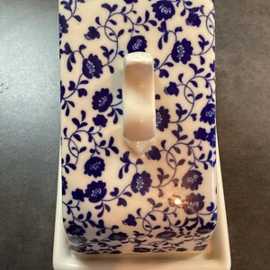 May include: A rectangular ceramic butter dish featuring a white base and a removable lid decorated with a blue floral pattern on a white background. The lid includes a central white handle and a small circular hole at the top edge for ventilation.