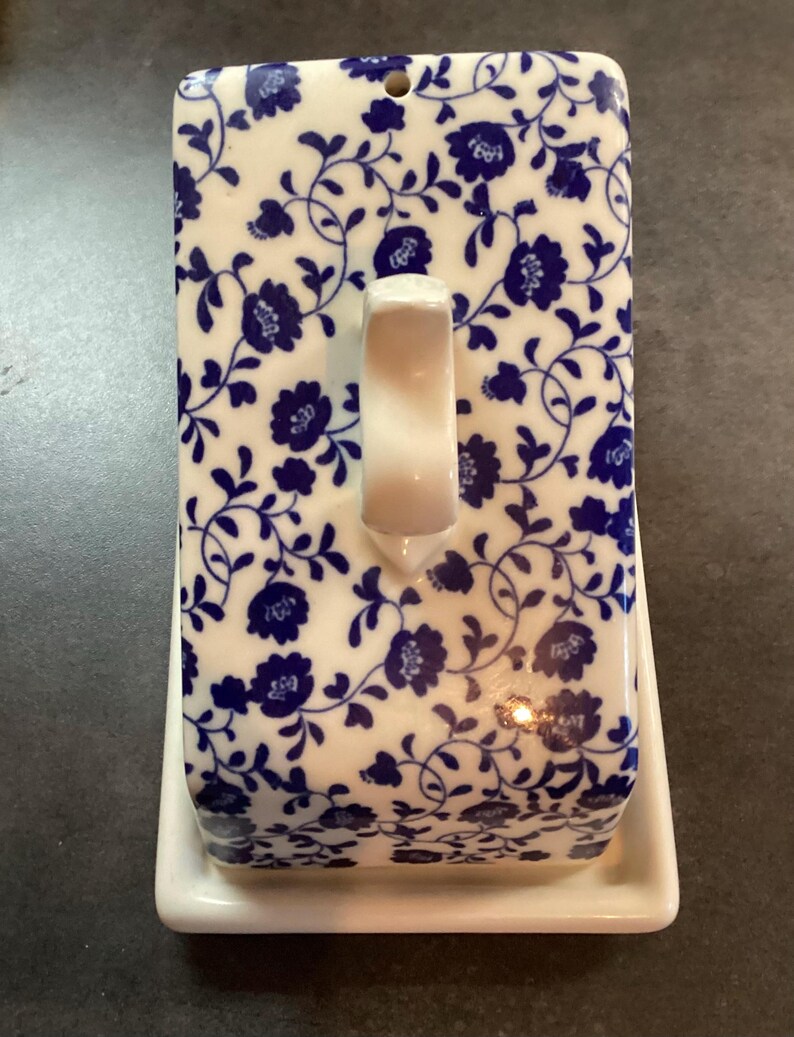 May include: A rectangular ceramic butter dish featuring a white base and a removable lid decorated with a blue floral pattern on a white background. The lid includes a central white handle and a small circular hole at the top edge for ventilation.