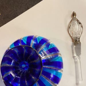 May include: A round glass perfume bottle featuring alternating vertical stripes of cobalt blue, turquoise, and clear glass with silver glitter accents. The bottle has a dark blue circular stopper and a separate clear glass applicator wand.