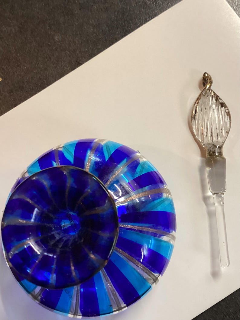 May include: A round glass perfume bottle featuring alternating vertical stripes of cobalt blue, turquoise, and clear glass with silver glitter accents. The bottle has a dark blue circular stopper and a separate clear glass applicator wand.