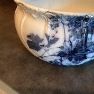 Pode incluir: Uma tigela de cerâmica cor de creme com um padrão de transferência azul escuro de folhas e cachos de uva. A tigela tem um design exterior canelado e recortado com desgaste visível e lascas ao longo da borda superior, mostrando o material base.