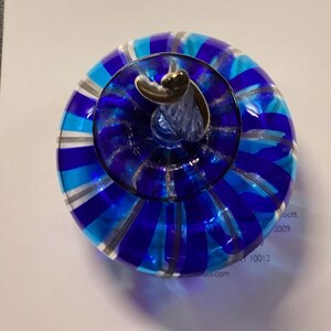 May include: A round glass paperweight featuring alternating vertical stripes of royal blue, light blue, and clear glass. The top centre has a small, clear glass stem-like protrusion with a metallic silver-coloured accent at the very peak.