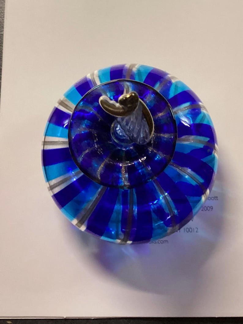 May include: A round glass paperweight featuring alternating vertical stripes of royal blue, light blue, and clear glass. The top centre has a small, clear glass stem-like protrusion with a metallic silver-coloured accent at the very peak.
