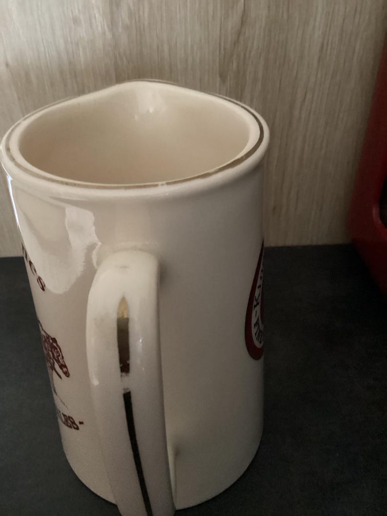 Op de afbeelding: Cr&egrave;mekleurige keramische mok met gouden rand en handvataccenten. De mok heeft een bedrukt ontwerp in bruin en rood, inclusief tekst en een afbeelding. De handgreep is gebogen en de mok heeft een brede opening.